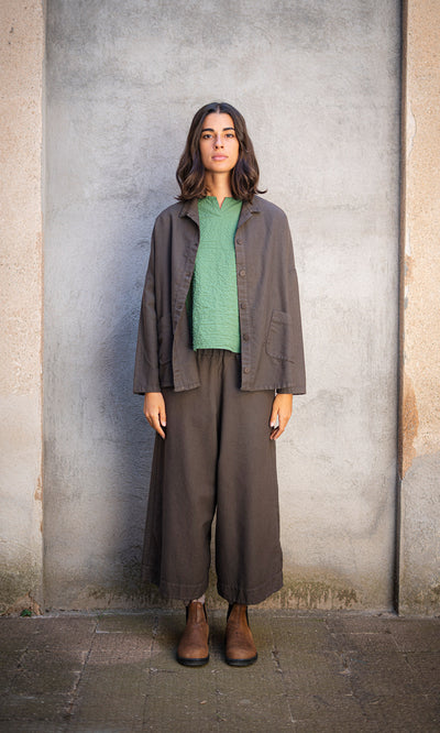 Plümo  Person with shoulder-length hair stands against a gray wall, wearing Otto Trousers and workwear boots.