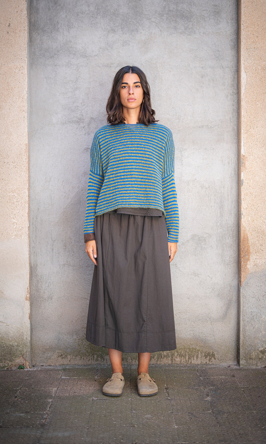 Plümo  Woman with shoulder-length brown hair in the Gracie Jumper and a long skirt, standing by a gray wall.