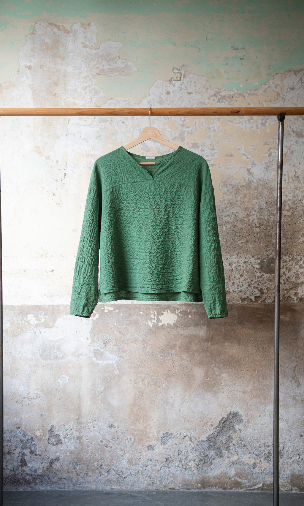 Plümo  The Verde Blouse, a green textured cotton top with a split neckline, hangs on a wooden hanger by a rustic wall.