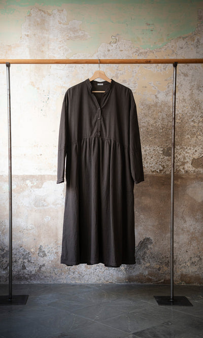 Plümo  The Marisol Dress - Truffle, a cotton long-sleeve style, hangs on a wooden hanger against a textured wall.