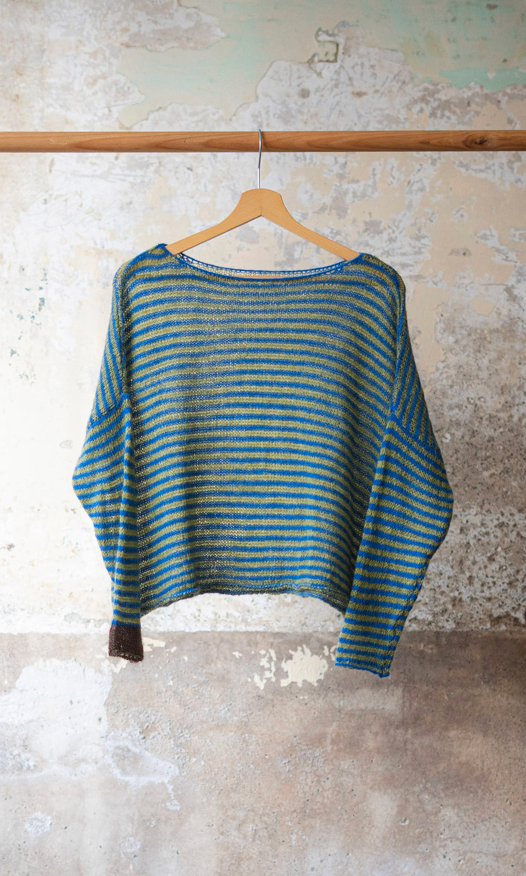 Plümo  The Gracie Jumper, a blue and green striped boat-neck sweater, hangs on a wooden hanger by a weathered wall.