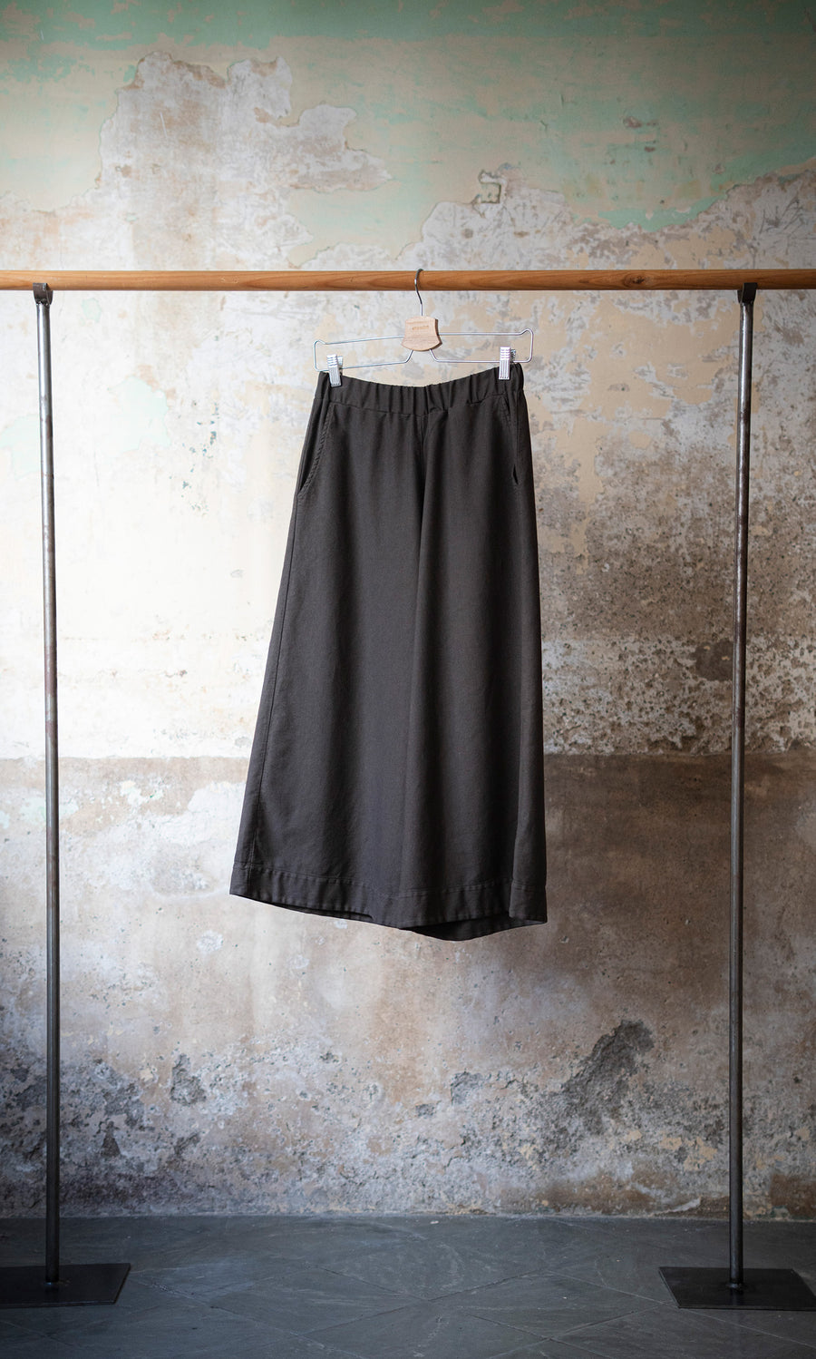 Plümo  Otto Trousers hang on a wooden rod, evoking classic painter’s workwear against a textured, distressed wall.