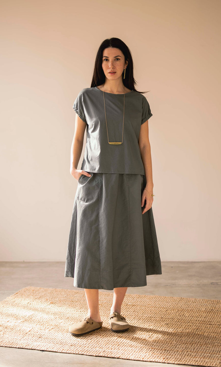 Plümo- Woman wearing a loose gray top and Marian Skirt, long necklace, beige shoes, standing on a woven rug indoors.
