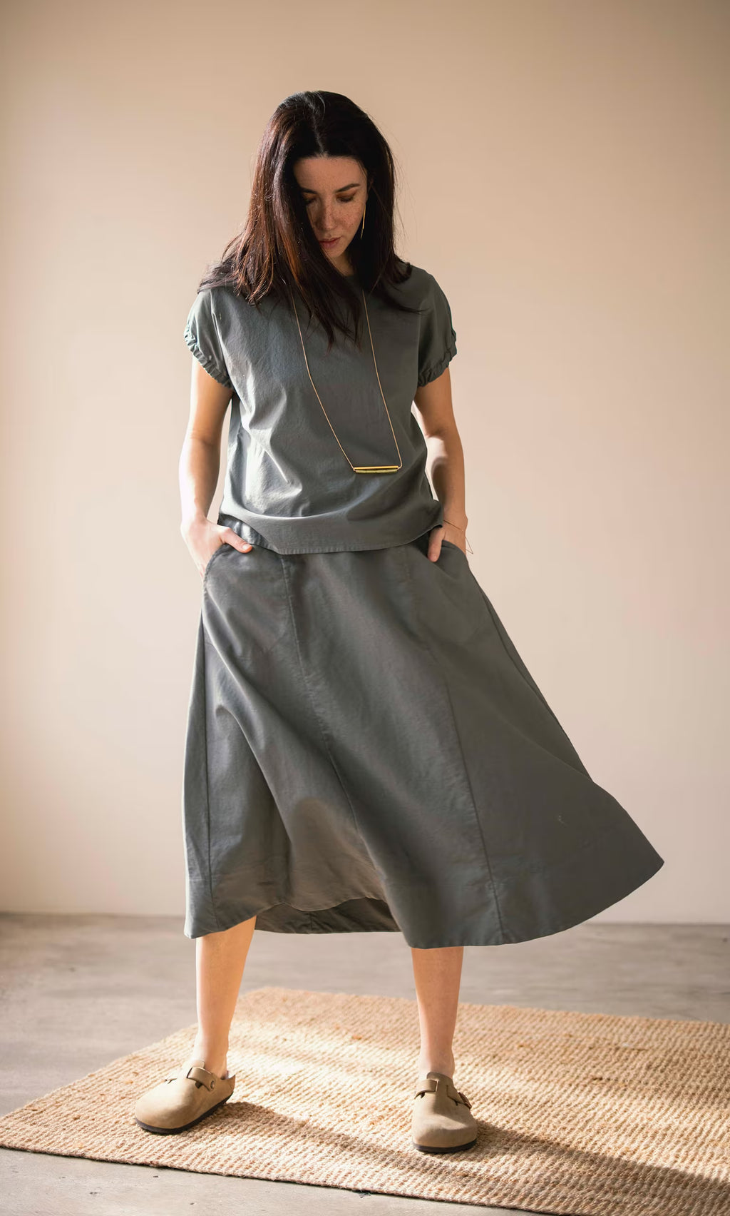 Plümo- Woman in a loose gray top and Marian Skirt stands on a woven rug, looking down with hands in pockets.