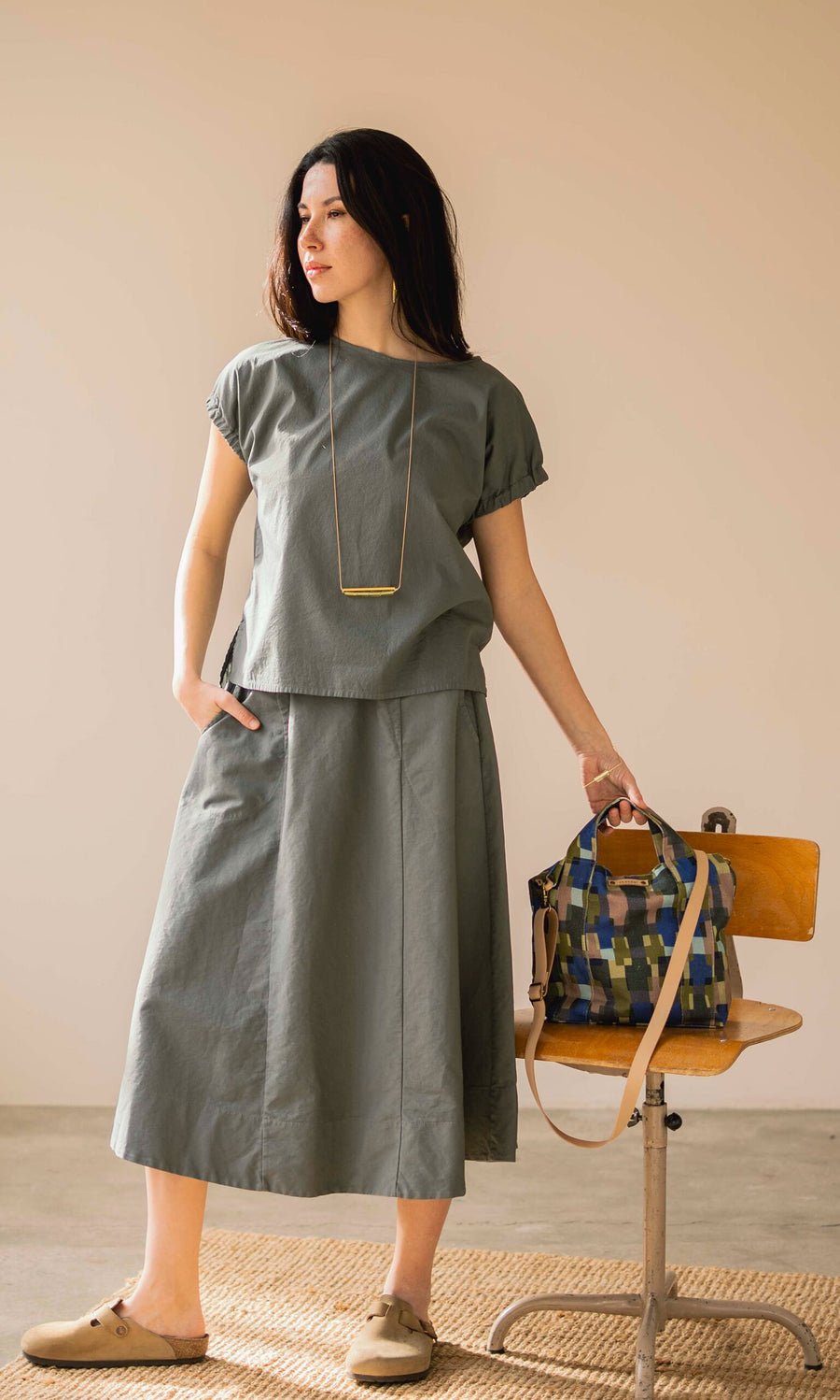 Plümo- Woman in a loose grey top and Marian Skirt stands by a chair, holding a patterned bag in natural light.
