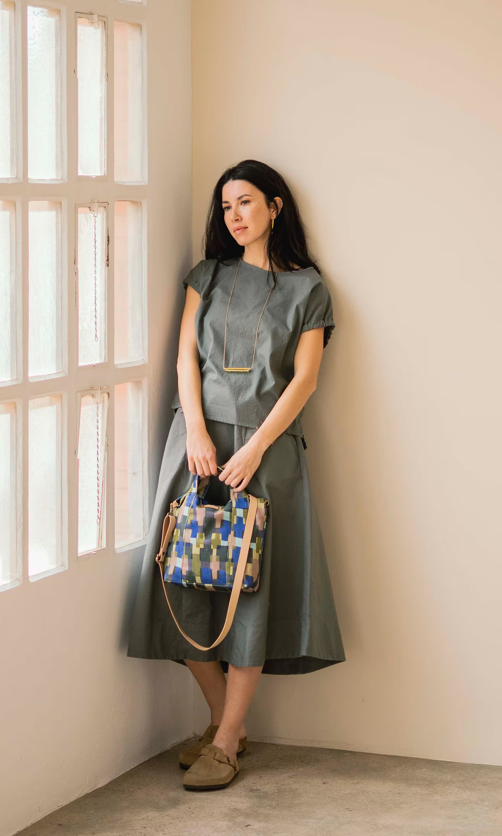 Plümo- Woman in a Felicity Blouse stands by a window, holding a colorful handbag and gazing outside thoughtfully.