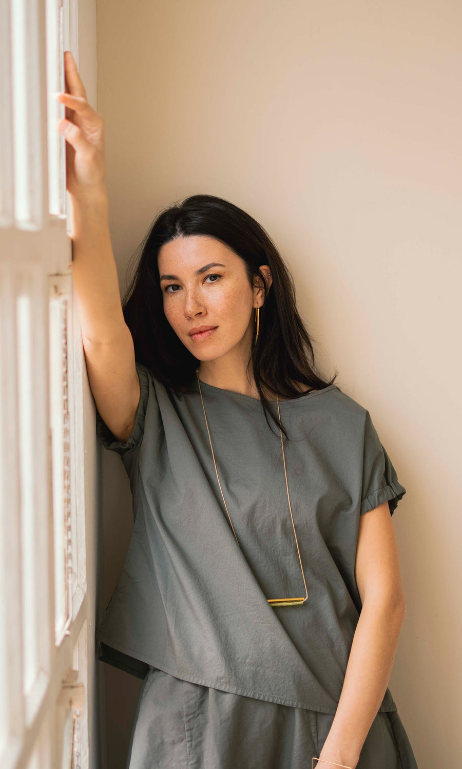 Plümo- Woman with long dark hair wears a loose gray top and the Lydia Necklace, leaning against a beige wall.