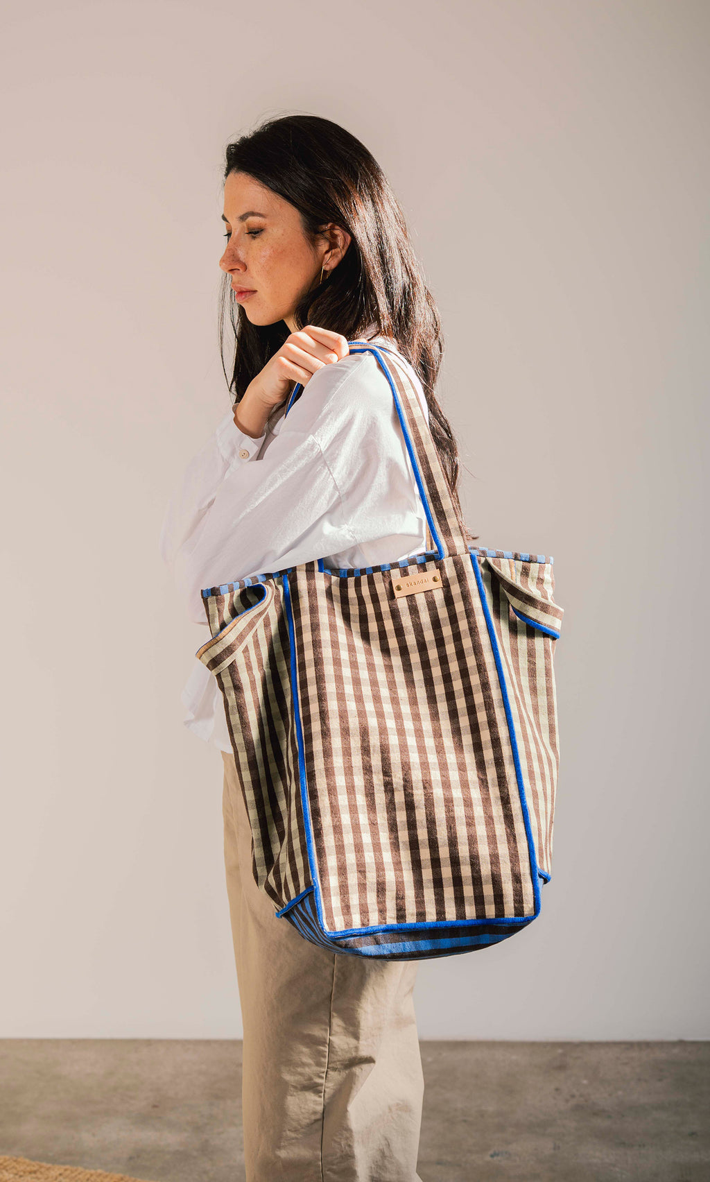 Plümo  Woman in white shirt carries the Stylist Bag, a spacious gingham tote with blue trim, over her shoulder.