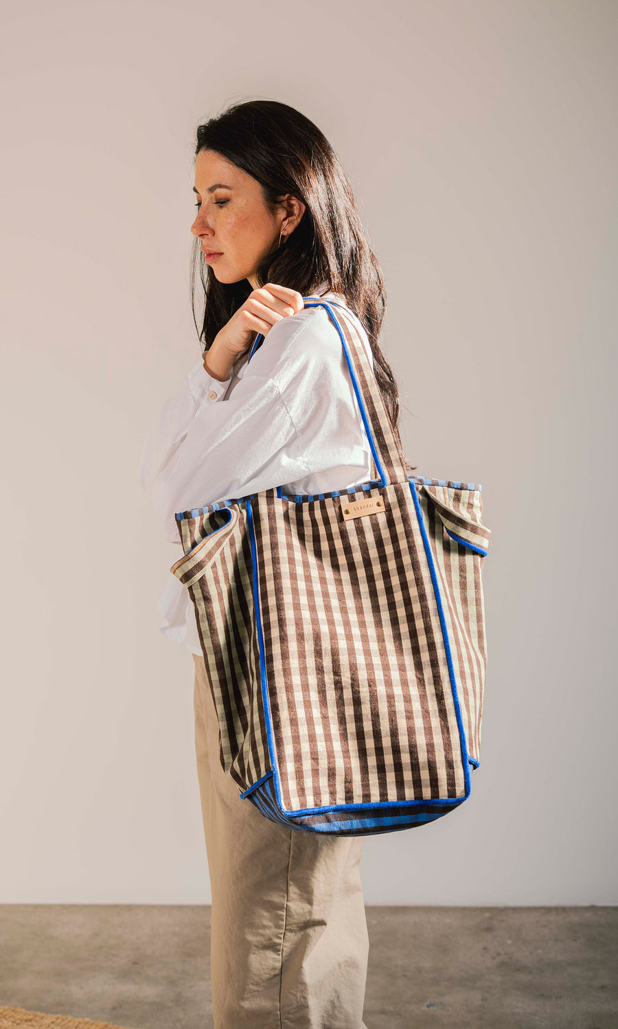 Plümo  Woman in white shirt carries the Stylist Bag, a spacious gingham tote with blue trim, over her shoulder.