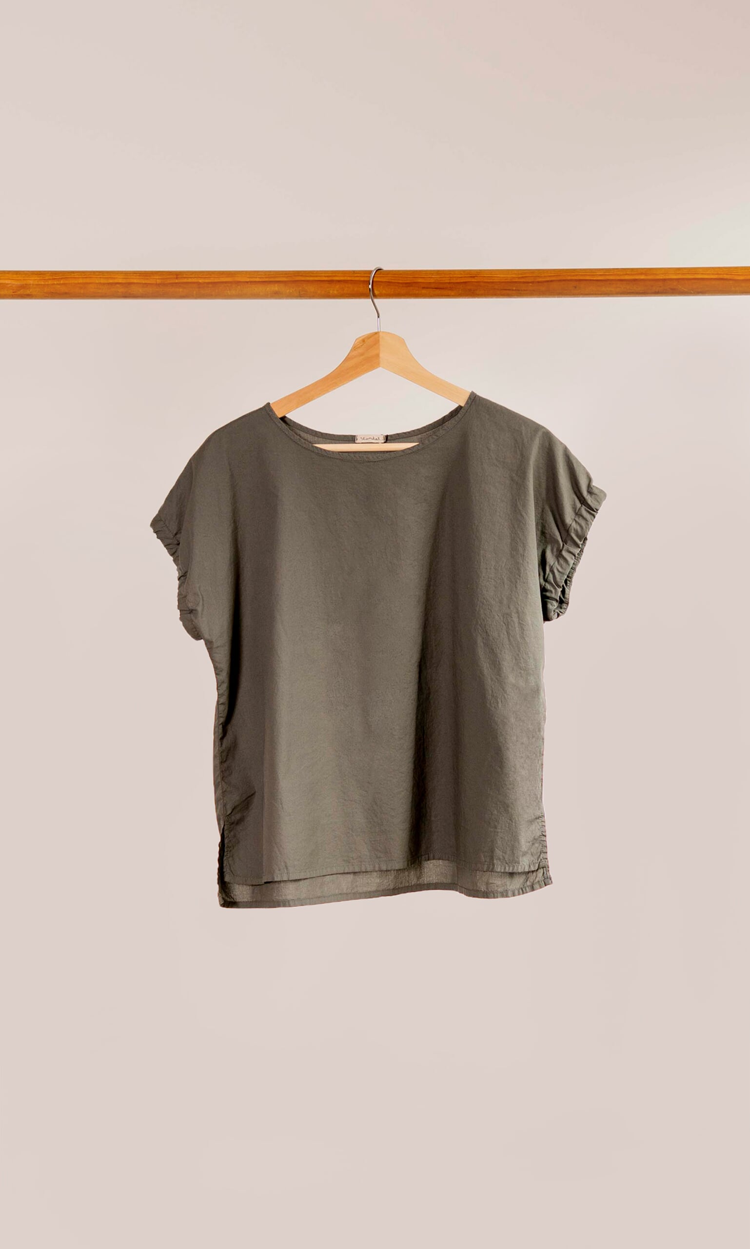 Plümo- The Felicity Blouse, a plain olive green top with cap sleeves, hangs on a wooden hanger against a light background.