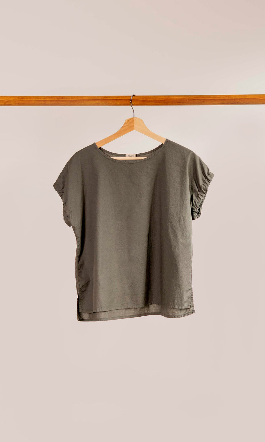 Plümo- The Felicity Blouse, a plain olive green top with cap sleeves, hangs on a wooden hanger against a light background.