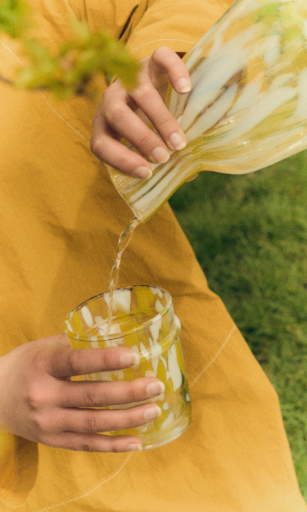 Plümo- Someone pours water from a Speckled Drinking Glass - Yellow into a matching glass outdoors.
