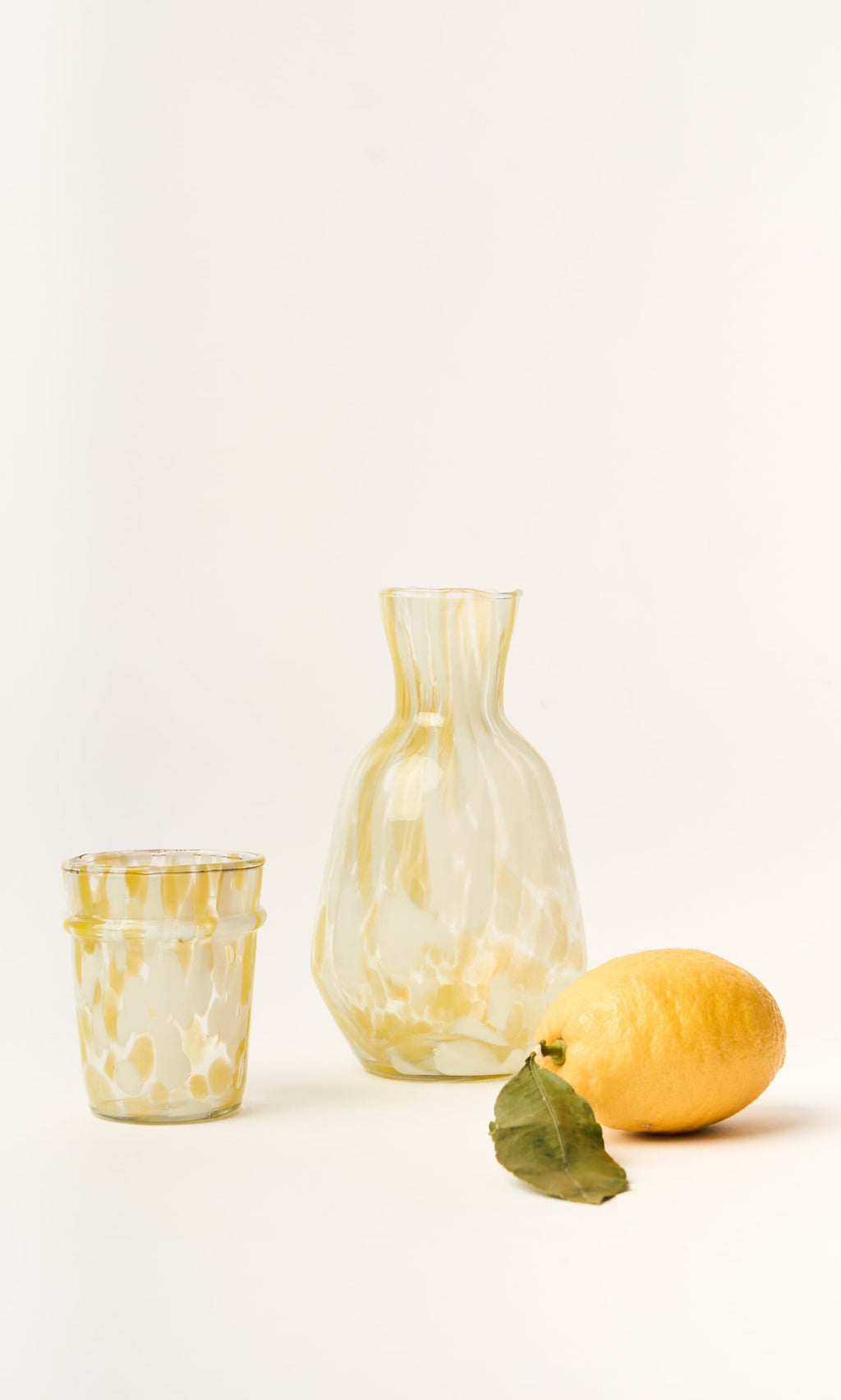 Plümo- The Speckled Drinking Glass - Yellow, handmade in Firozabad, features yellow marbling and sits beside a lemon and leaf.