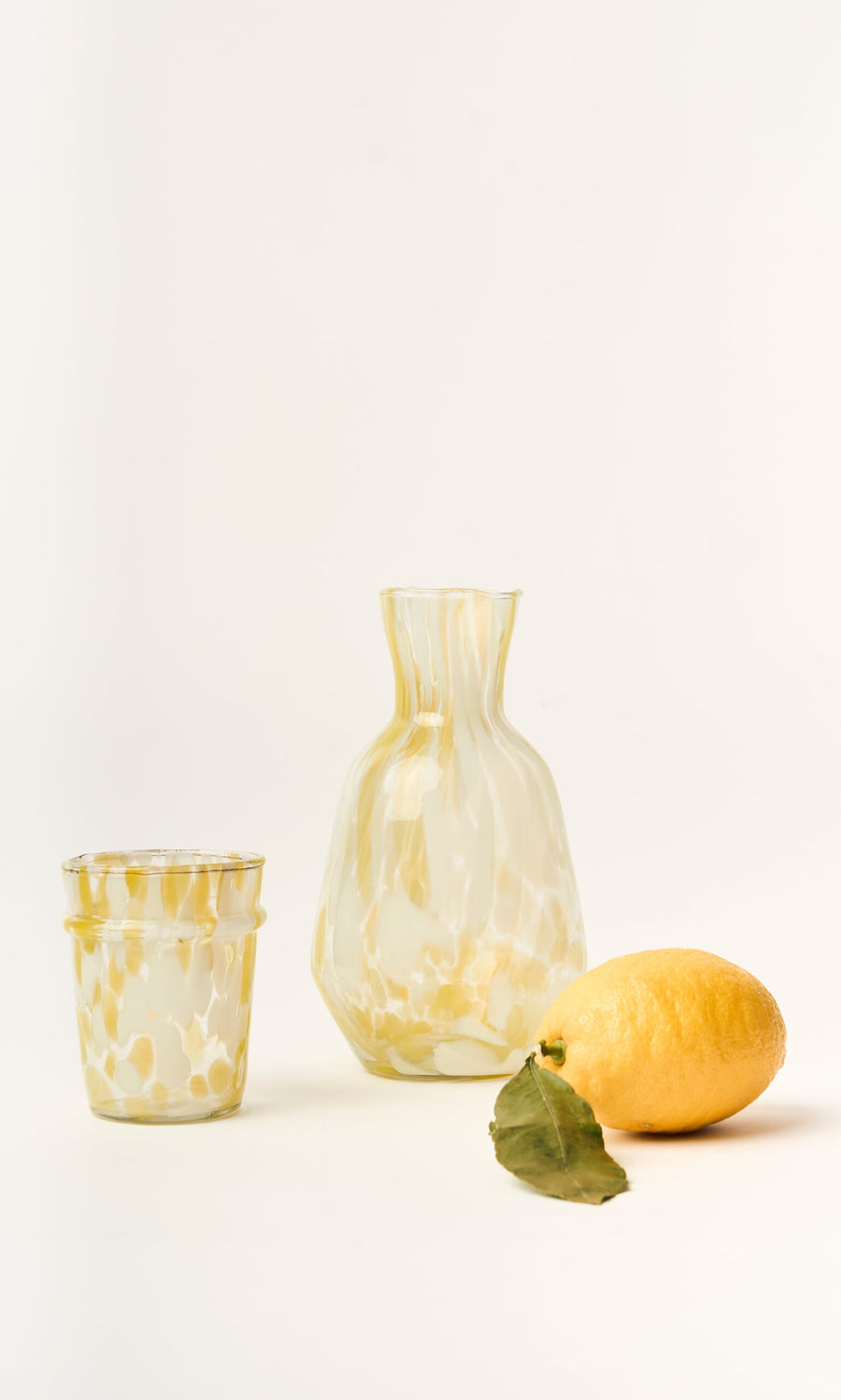 Plümo- The Speckled Drinking Glass - Yellow, handmade in Firozabad, features yellow marbling and sits beside a lemon and leaf.