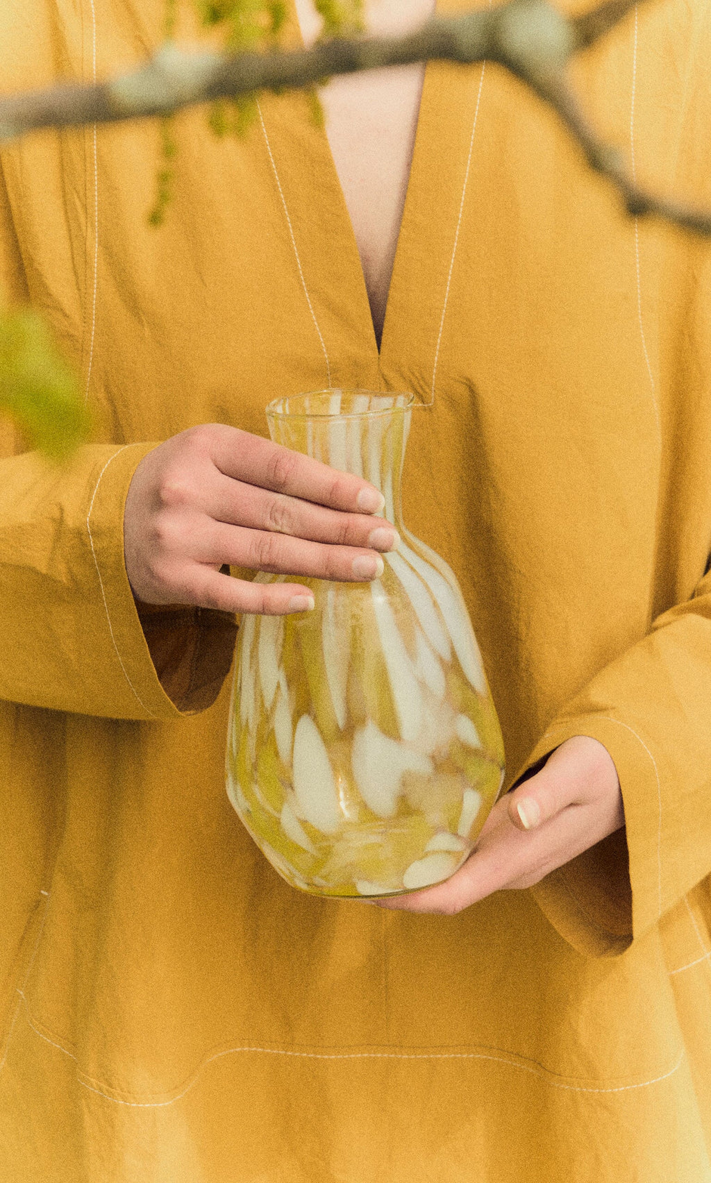 Plümo- Person in yellow holds a Speckled Glass Jug - Yellow, with branches visible in the foreground.