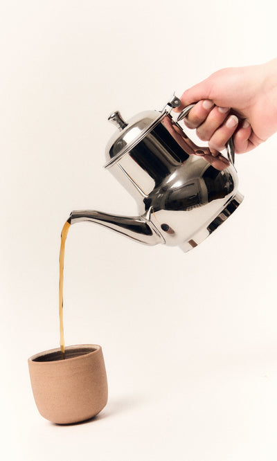 Plümo- A hand pours tea from a Steel Teapot with an industrial finish into a brown ceramic cup on a white background.