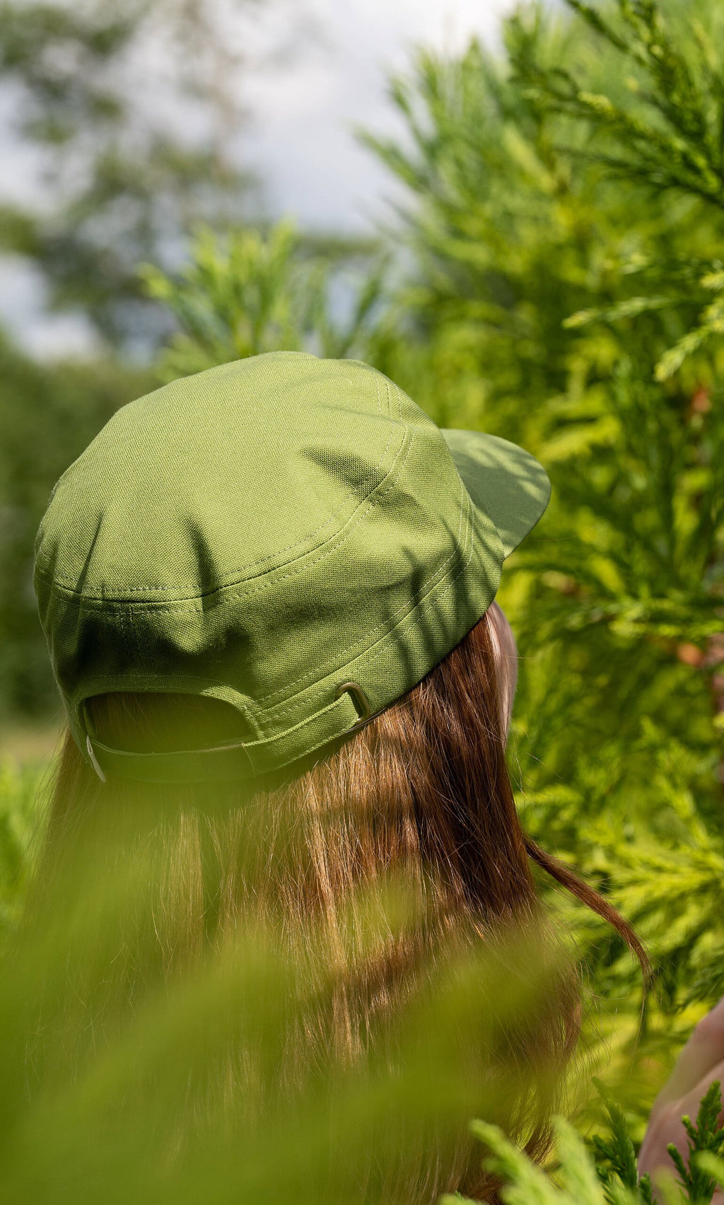 Plümo- Person with long brown hair in a green Tsuba Cap stands among bright green foliage, facing away.