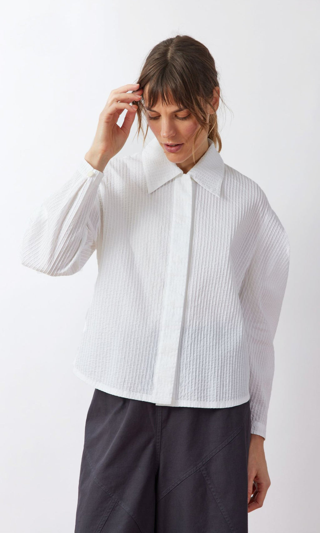 Plümo- Woman in a Ting Blouse with bishop sleeves and dark pants stands against a white background, looking down.
