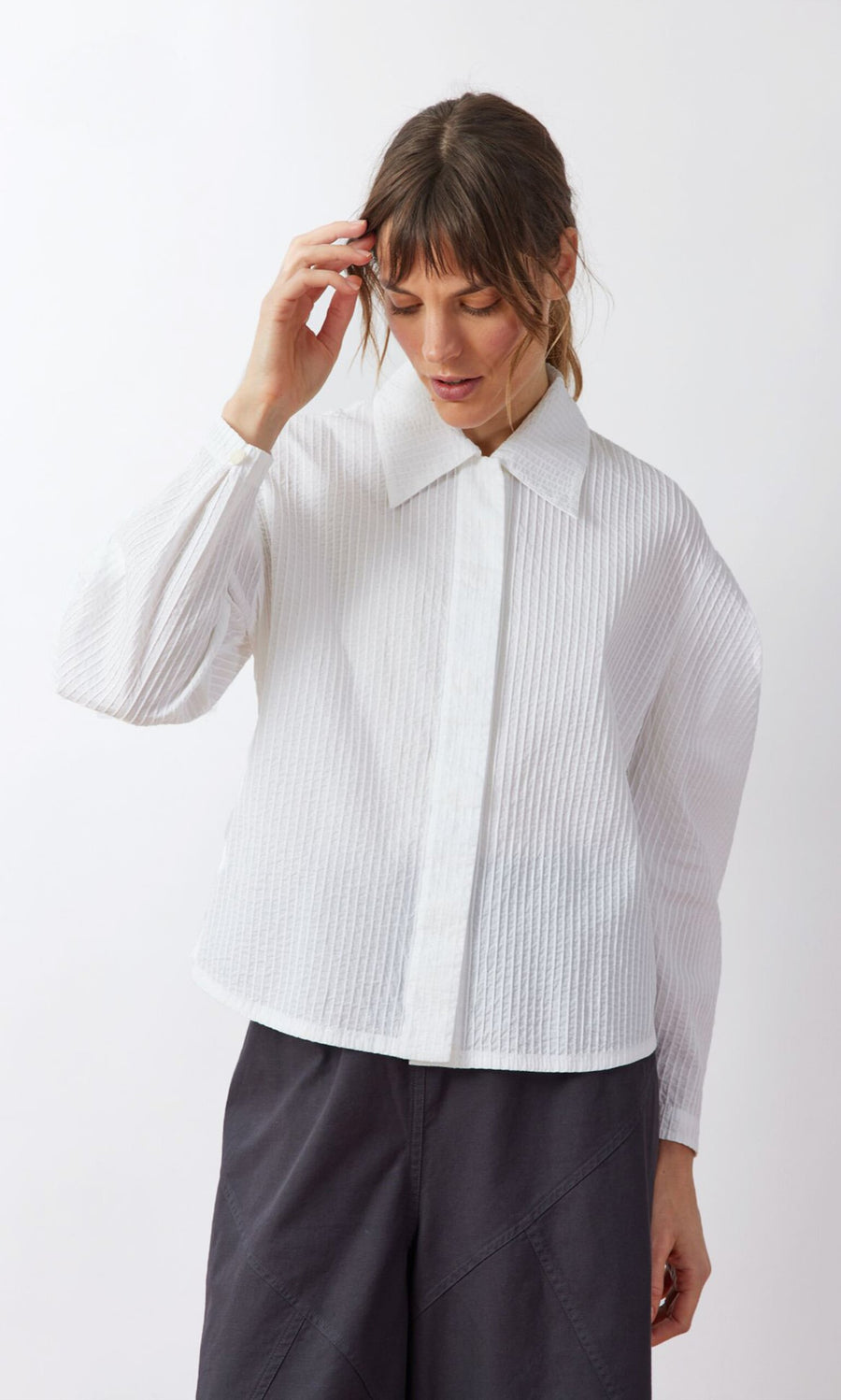 Plümo- Woman in a Ting Blouse with bishop sleeves and dark pants stands against a white background, looking down.