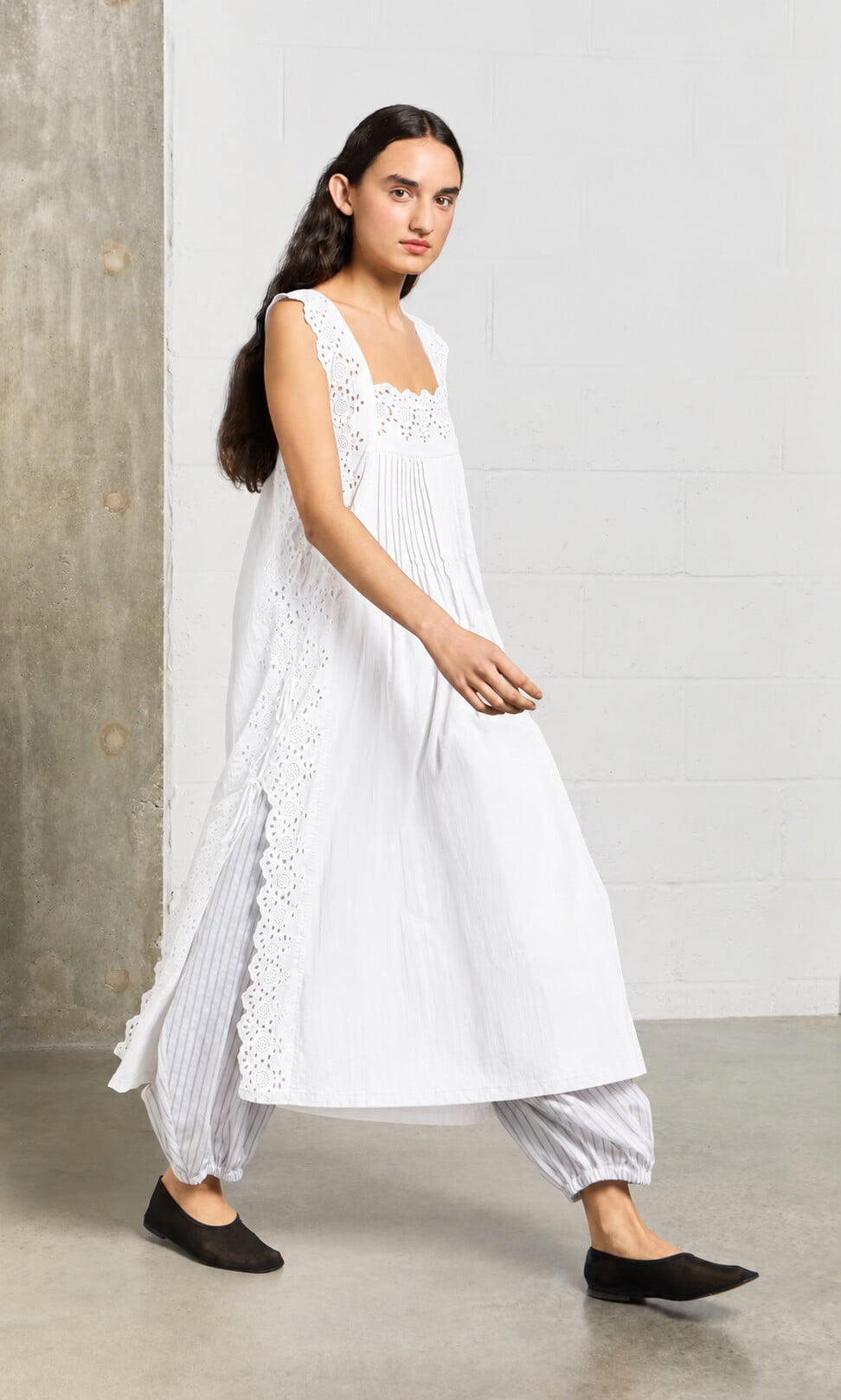 Plümo- A woman in a Vintage Lace Dress - White walks indoors against a concrete wall.