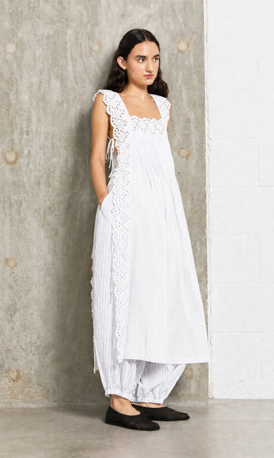 Plümo- Woman in a Vintage Lace Dress - White over striped pants, standing against a concrete wall.