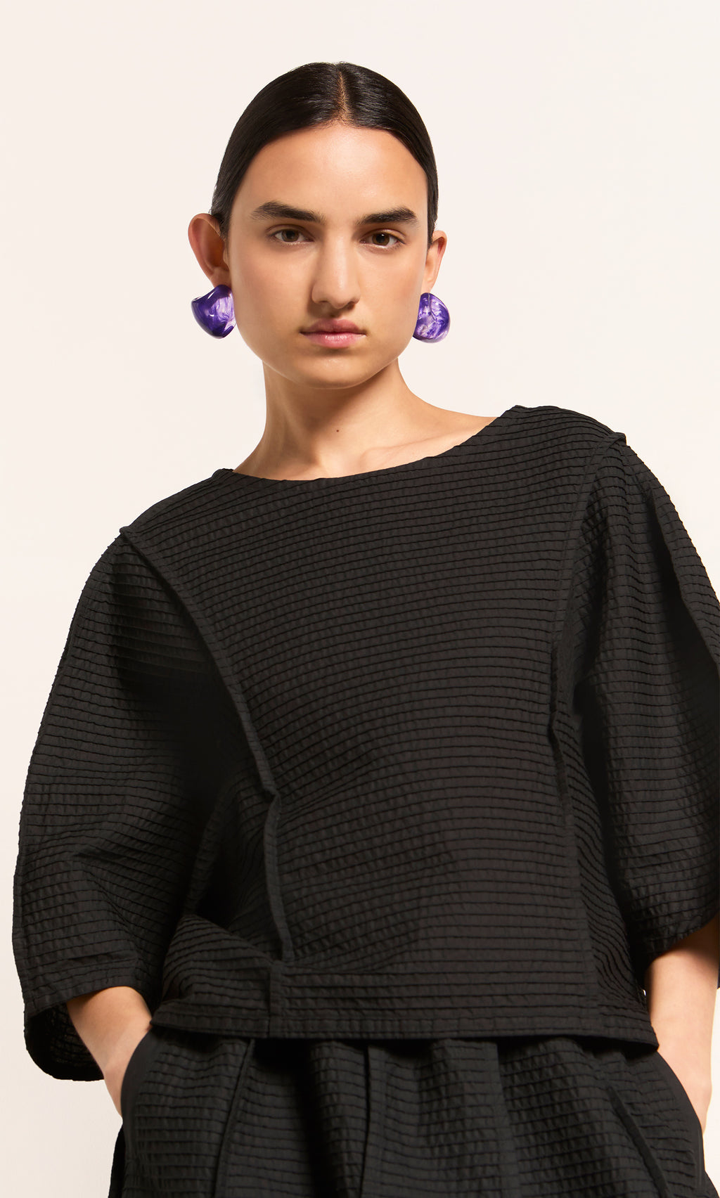 Plümo  Person in a textured black top, black pants, and bold Viola Earrings, standing against a plain background.