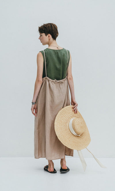 Plümo- Model in the Violet Dress, a sleeveless green linen slip with adjustable fit, holds a straw hat against a white background.