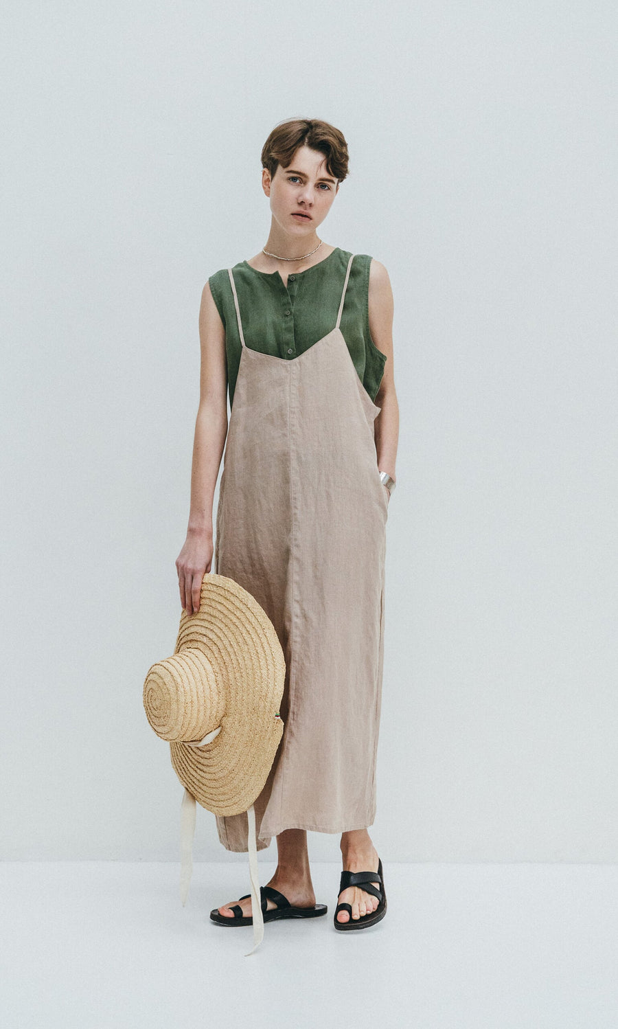 Plümo- Person wearing the Violet Dress, a sleeveless green linen slip with deep V-neck and adjustable fit, holding a straw hat.