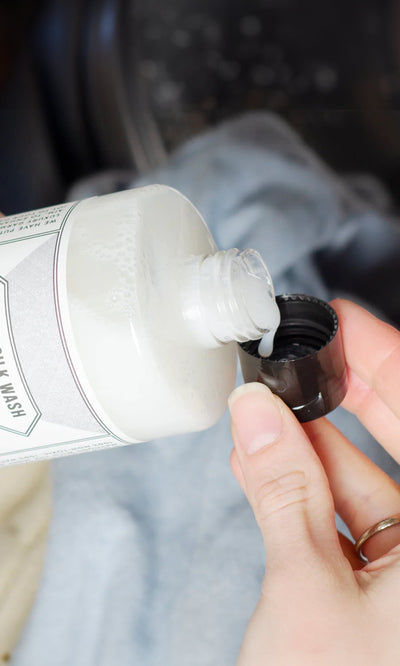 Plümo- A person pours Wool & Silk Detergent into a black cap, with clothes visible in the background.