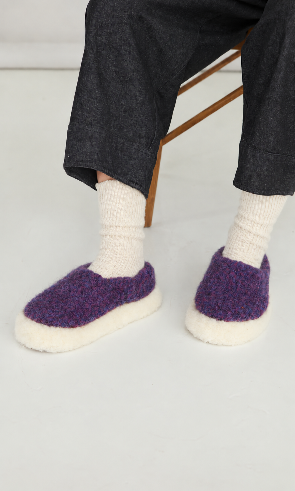 Plümo  Person in white socks and Plum Wool Slippers with non-slip soles, sitting on a wooden chair.