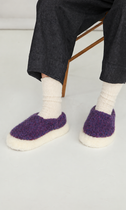 Plümo  Person in white socks and Plum Wool Slippers with non-slip soles, sitting on a wooden chair.