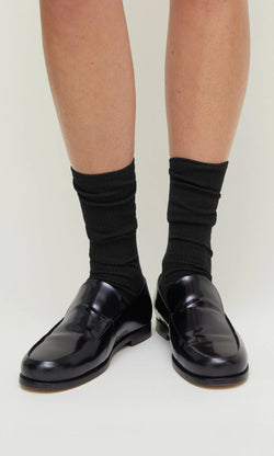 Plümo- Person wearing Black Rib Socks and black loafers, standing on a light-colored surface.
