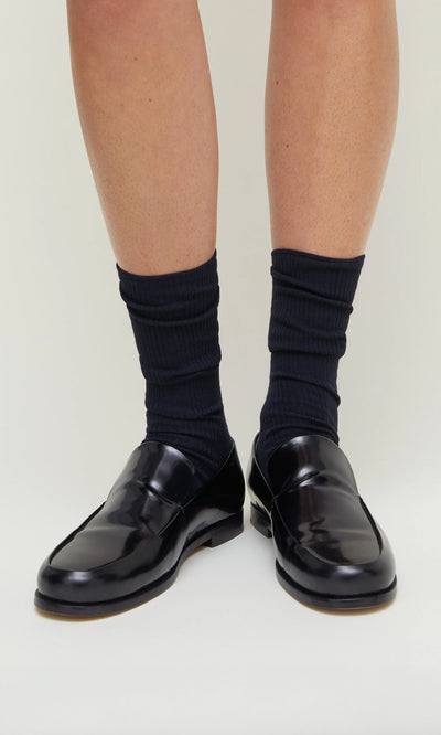 Plümo- Navy rib socks worn with shiny black loafers on a person's feet, standing on a light background.