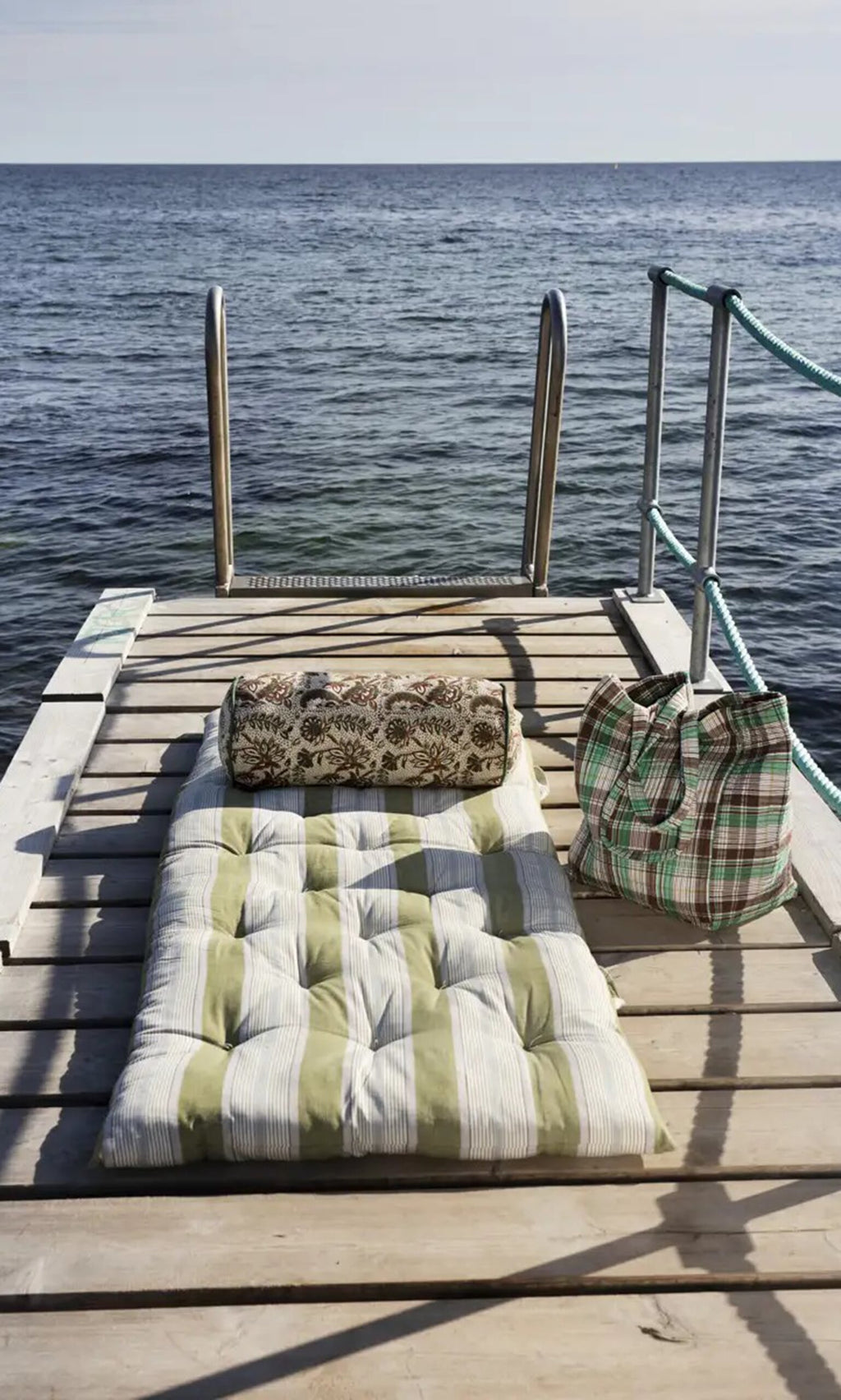 Plümo- A Printed Bolster Cushion with floral vines rests on a wooden dock by the sea beside a plaid bag and metal ladder.