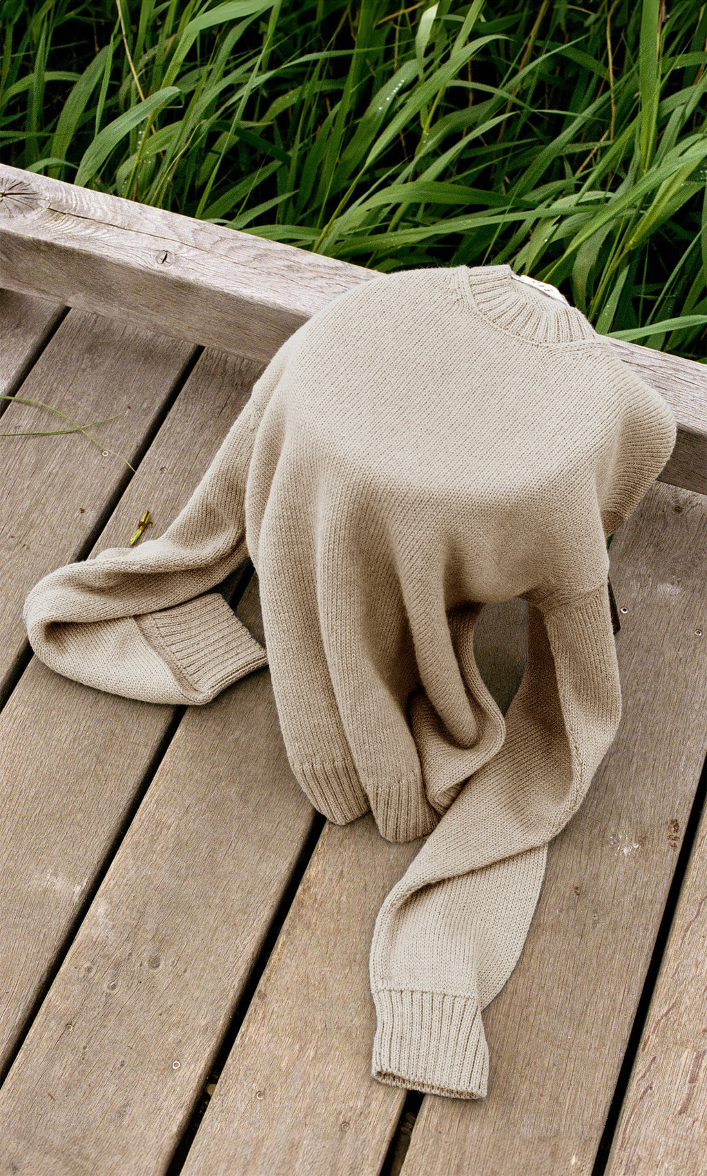 Plümo- The Christie Wool Jumper, a beige crewneck in a boyfriend fit, is draped on a wooden deck with green grass behind.