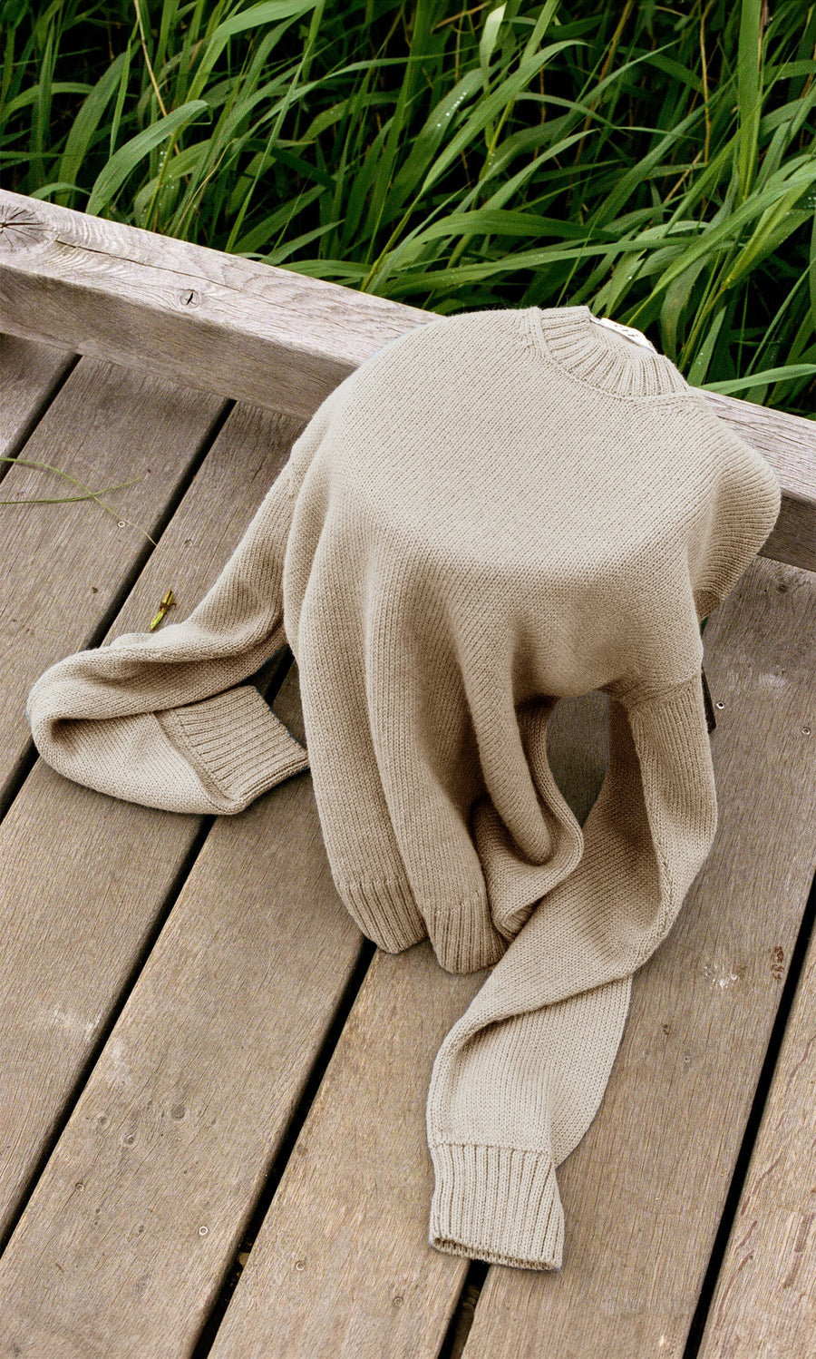 Plümo- The Christie Wool Jumper, a beige crewneck in a boyfriend fit, is draped on a wooden deck with green grass behind.