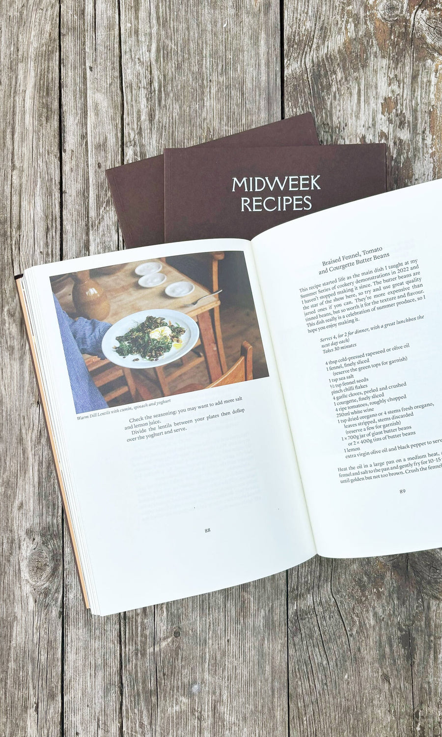 Plümo- Open recipe book on wooden table, with "Midweek Recipes By Jess Elliott Dennison" stacked underneath.