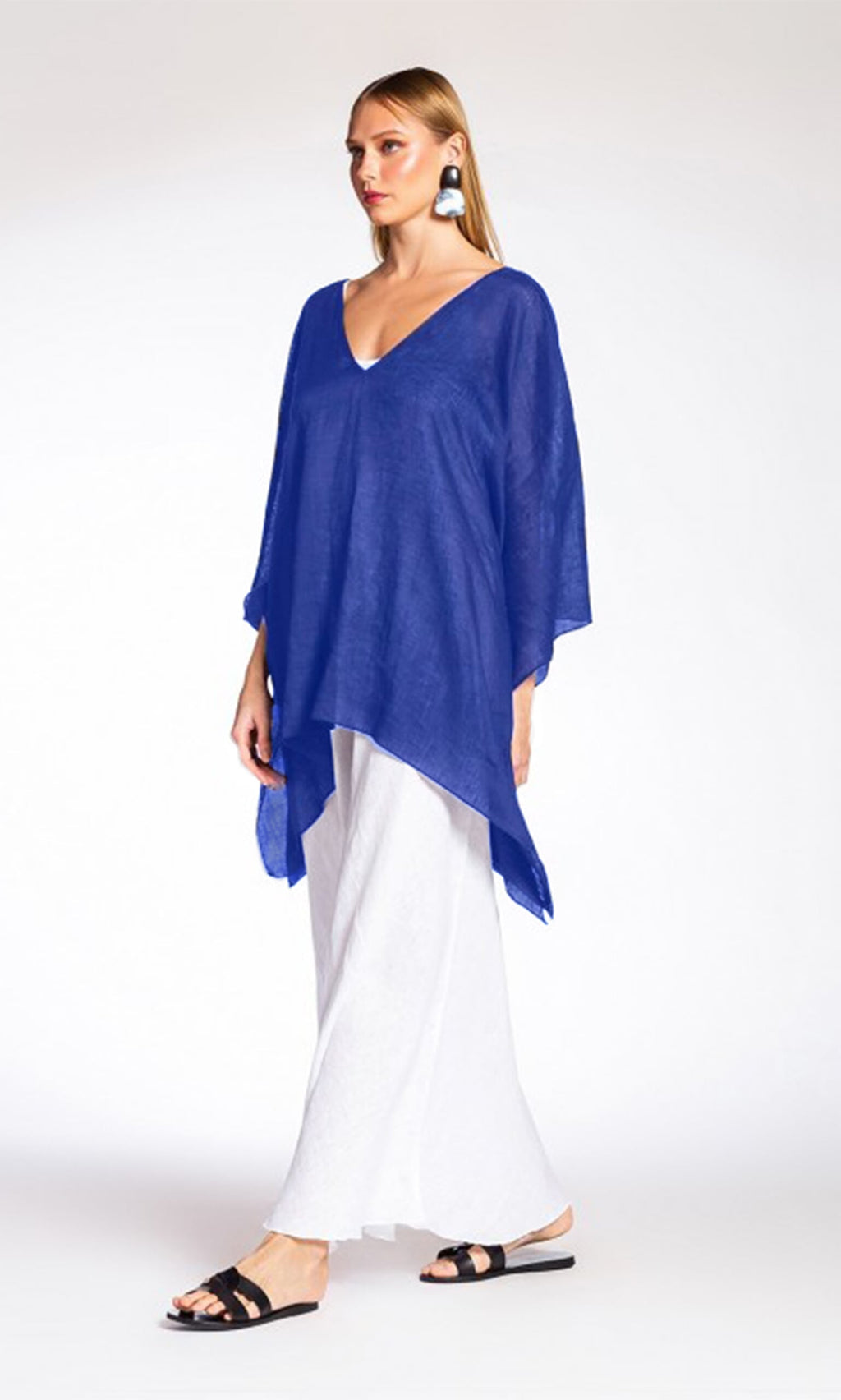 Plümo- Woman wearing the Drew Poncho, white wide-leg pants, black sandals, and white statement earrings.