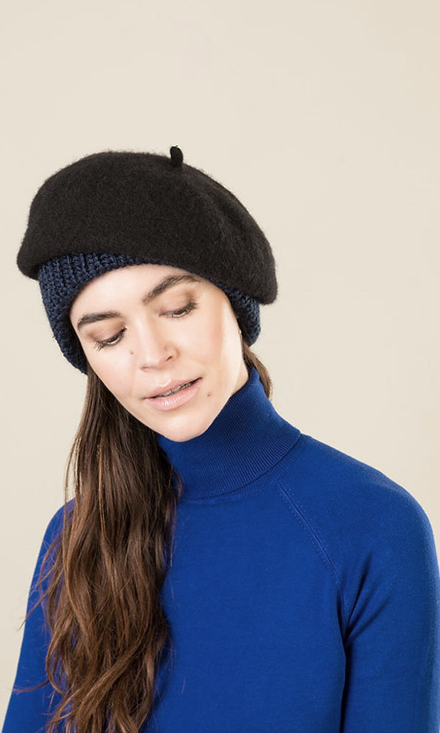 Plümo- Woman wears the Ashmere Beret and a blue turtleneck with navy knit panel, looking down against a neutral background.
