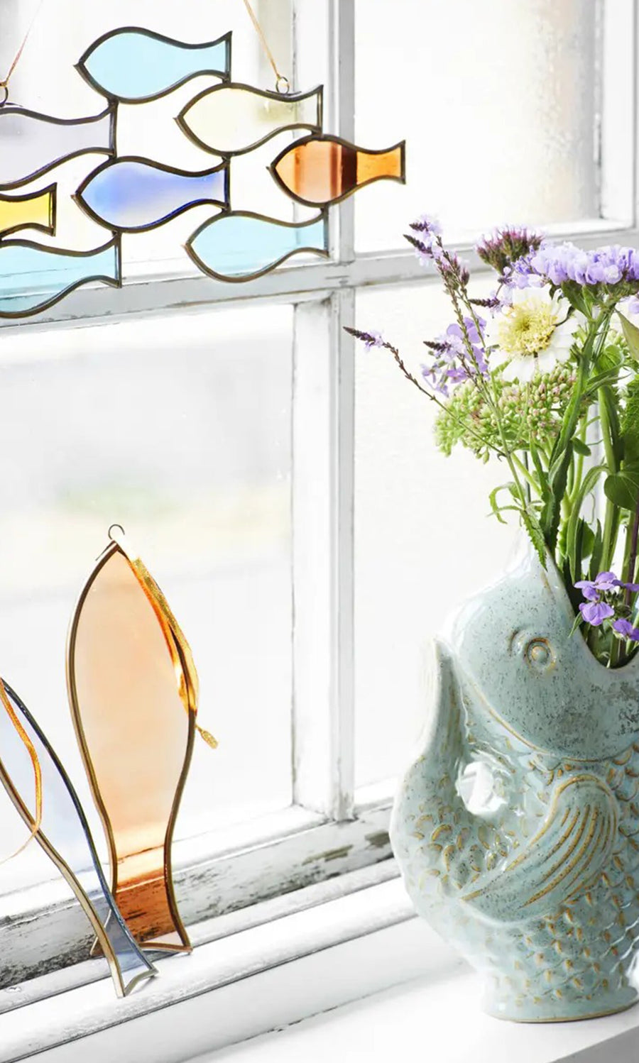 Plümo- The Shoal of Fish Mobile hangs in the window next to a ceramic vase with purple and white flowers.
