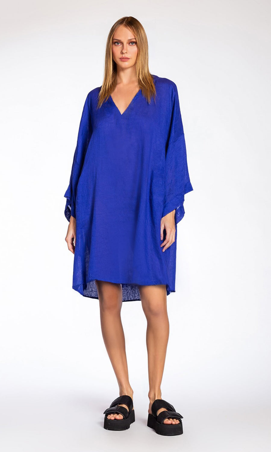 Plümo- Woman in a Luma Dress, a loose bright blue linen dress with a relaxed fit, and black platform sandals against white backdrop.