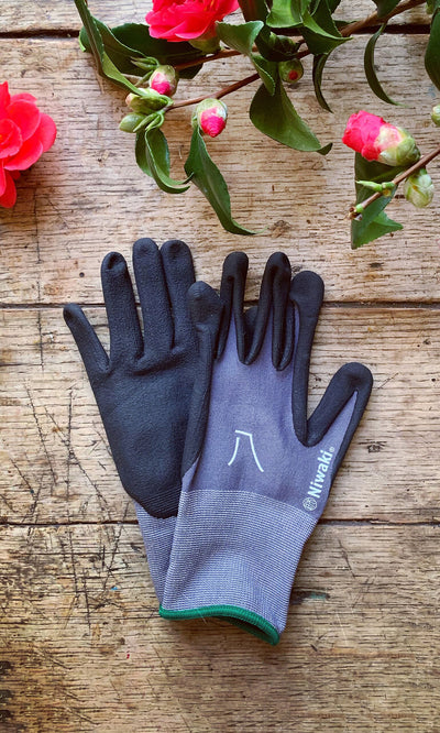 Plümo- A pair of black and gray Niwaki gardening gloves with nitrile coating on rustic wood, pink flowers in the corners.