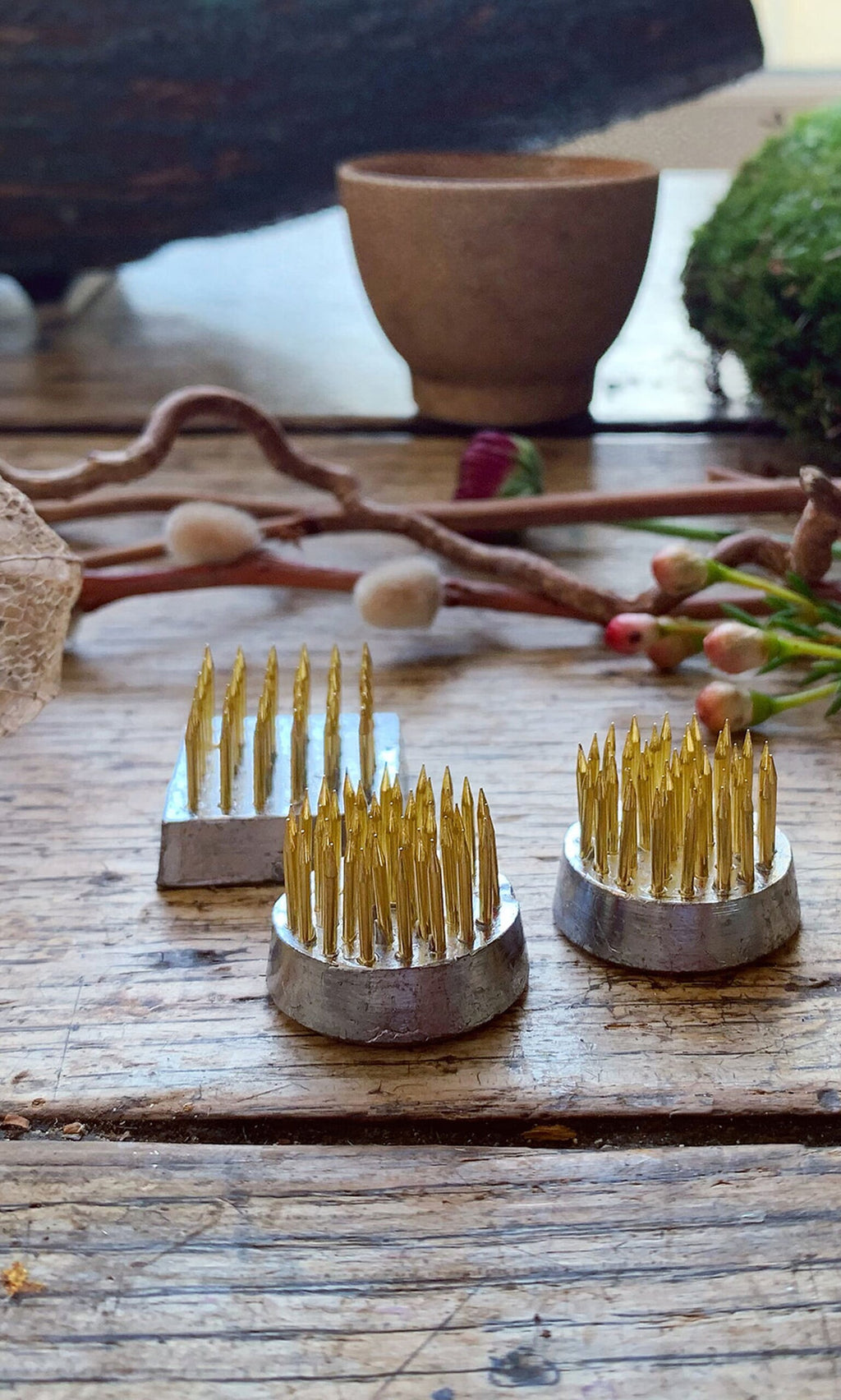 Plümo- Three Kenzan flower frogs, including a brass one, rest on a rustic wooden table with natural decor in the background.