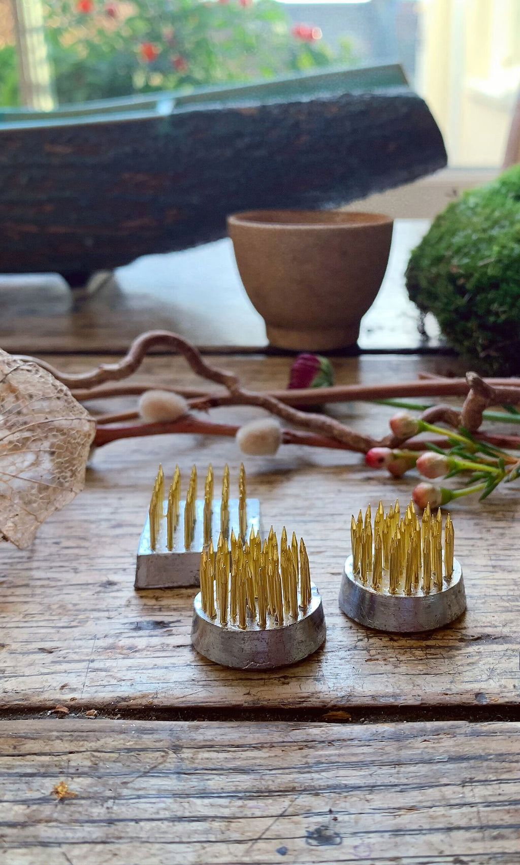 Plümo- Three Kenzan flower frogs with sharp gold pins rest on a rustic wooden table with natural objects in the background.
