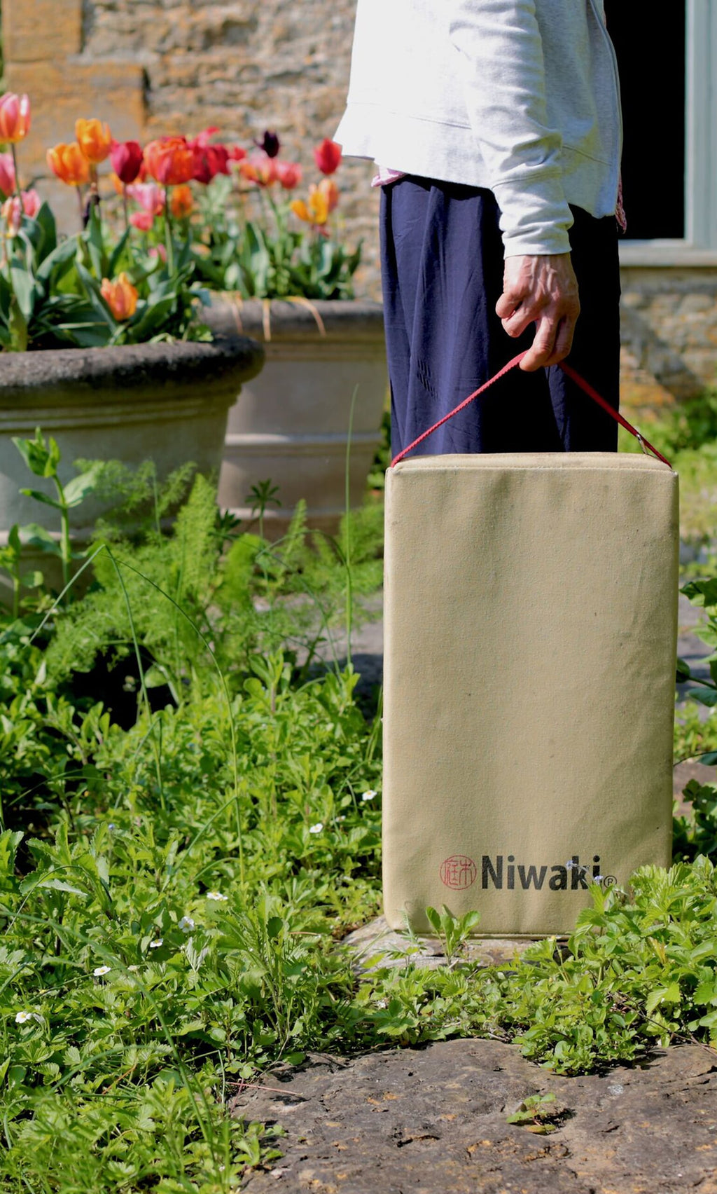 Plümo- Person holds a beige Niwaki Kneeler bag with a red handle, surrounded by flowers and greenery.