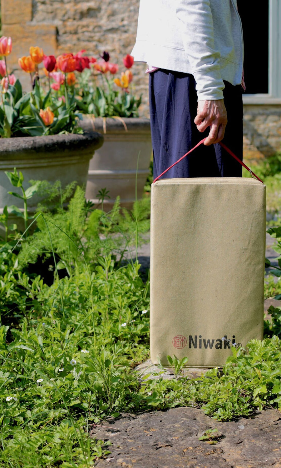 Plümo- Person holds a beige Niwaki Kneeler bag with a red handle, surrounded by flowers and greenery.