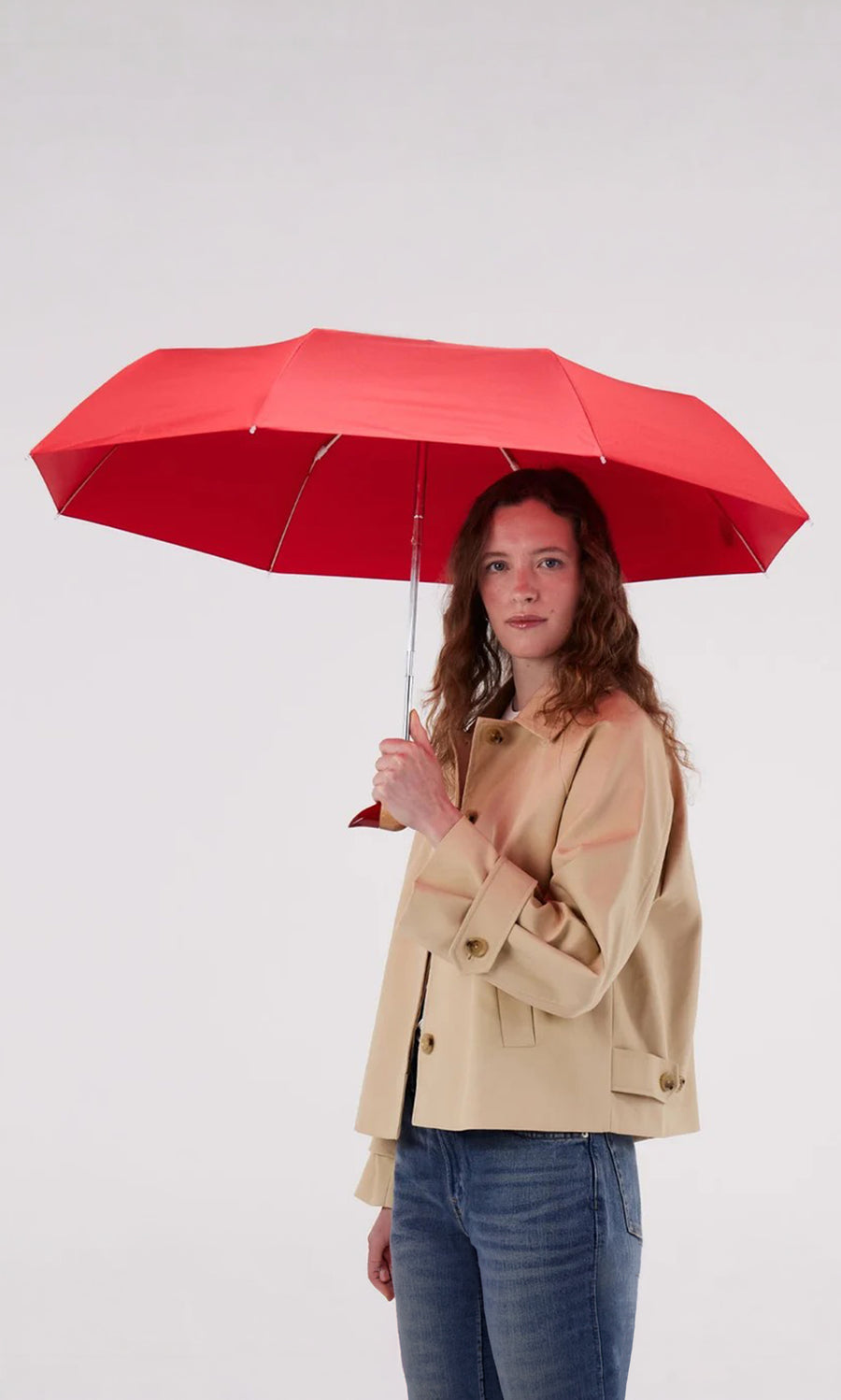 Plümo  Woman with wavy hair holds a red Duck Umbrella, wearing a beige jacket and blue jeans on a plain background.