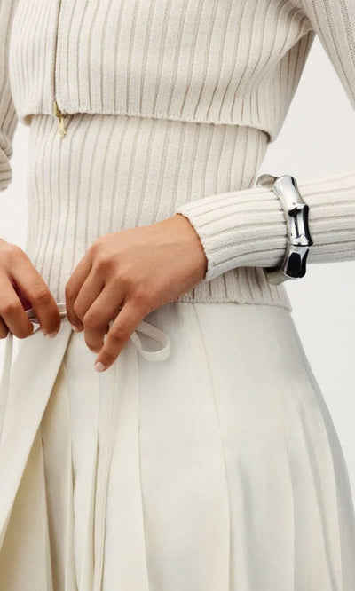 Plümo- Wearing a ribbed cream sweater and skirt, she ties a white ribbon with the Ridged Bangle on her wrist.