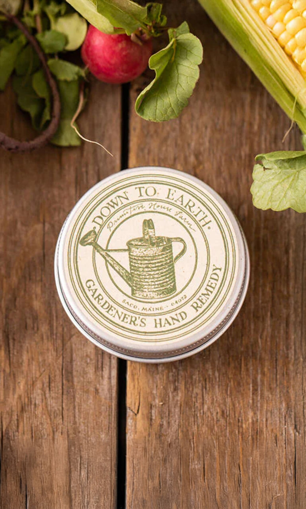 Plümo- A tin of Gardener's Hand Remedy balm rests on a wooden table with fresh vegetables nearby.