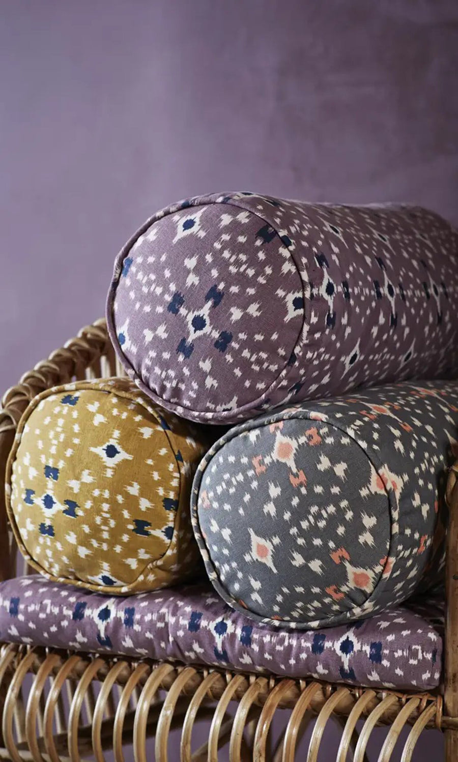 Plümo- A Printed Bolster Cushion - Grey is stacked on a wicker chair set against a purple wall.